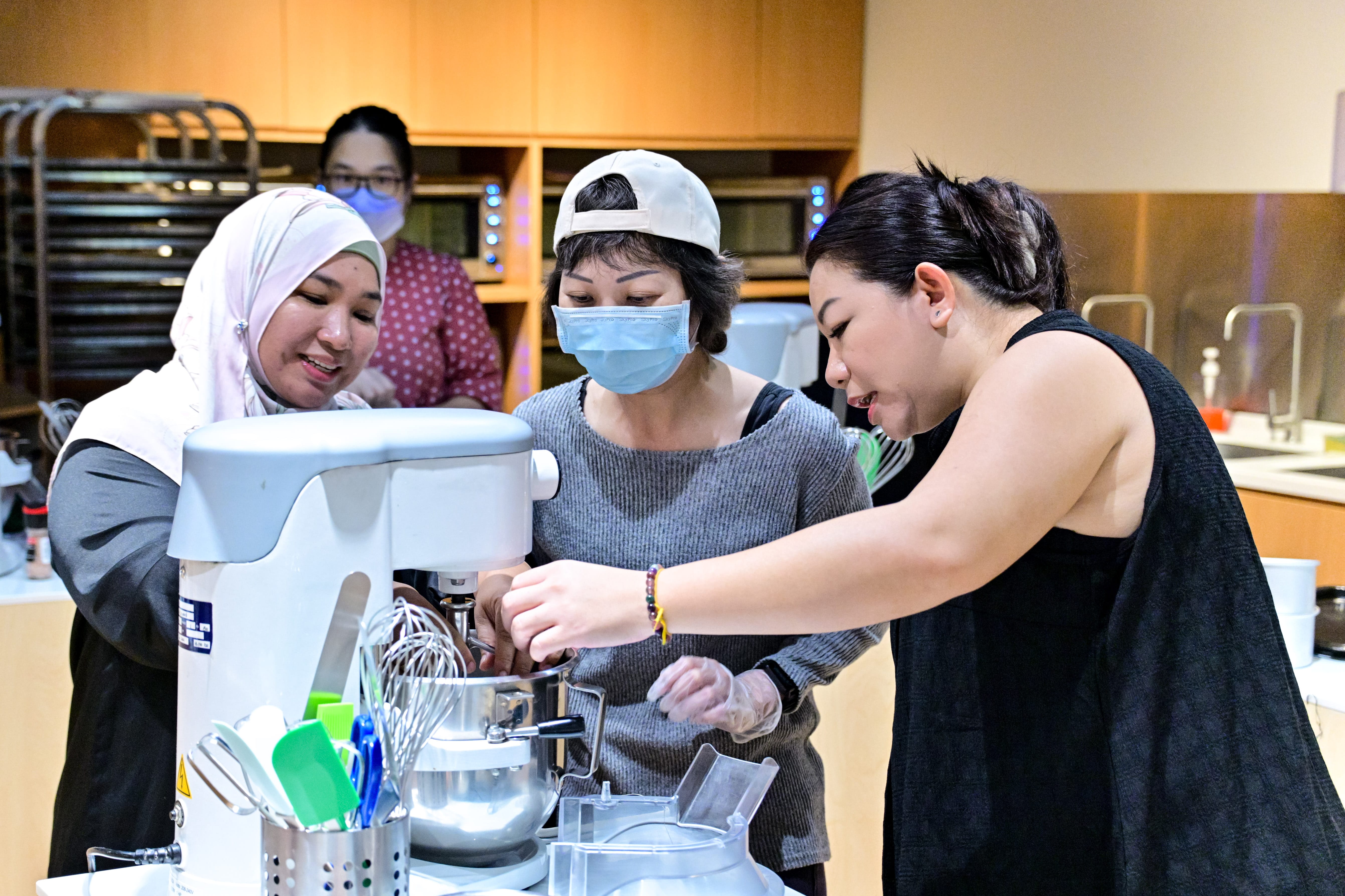 Three women gathered around a stand mixer, working together during a baking session.